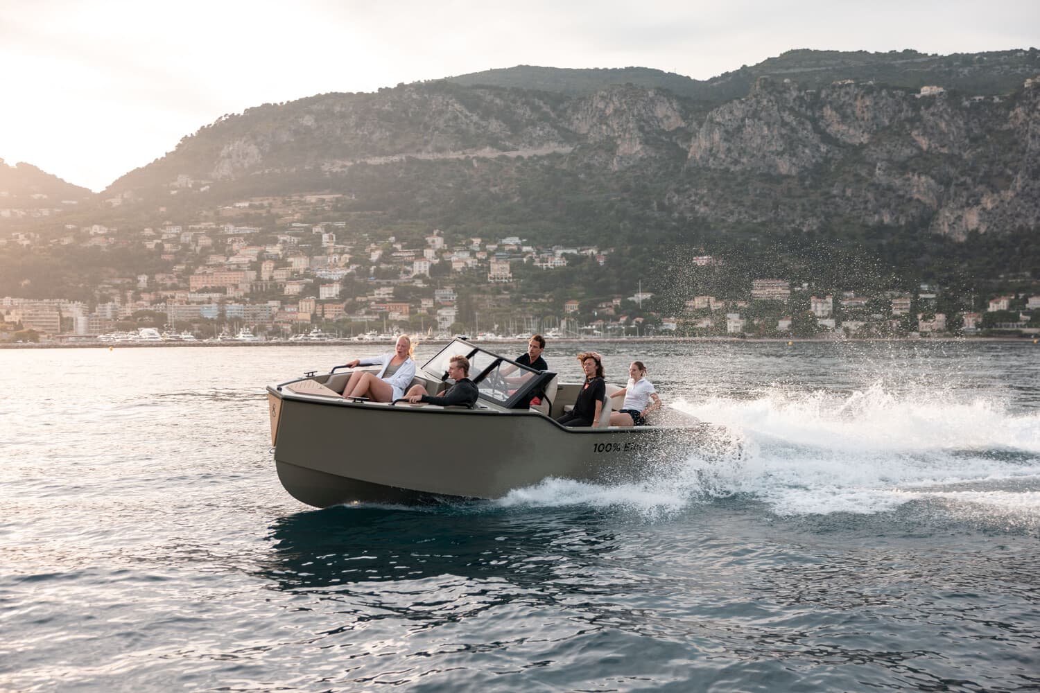 X Shore 1 motorboat cruising on coastal water with five passengers aboard, viewed from the side against a mountainous shoreline.