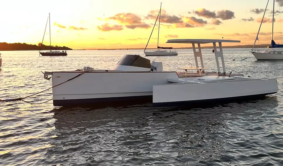 Novaluxe X-Wing electric boat floating on calm water at sunset, with sailboats and a distant shoreline in the background.