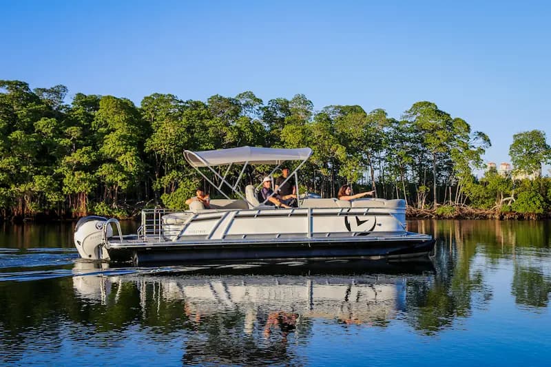 White Pontoon boat cruising on calm water with passengers aboard, canopy overhead, and mangrove-lined shore in the background. Vision marine branding on the side