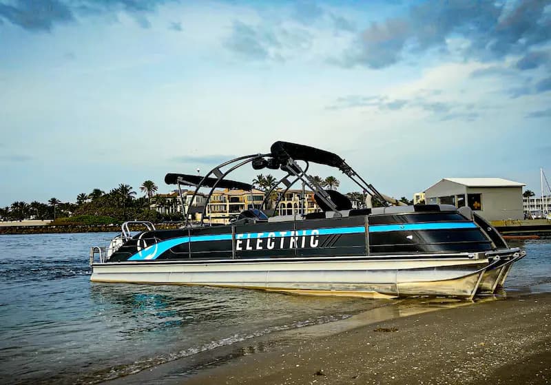 Vision Marine V30 electric pontoon boat beached at the water’s edge, with “Electric” branding and waterfront buildings in the background.