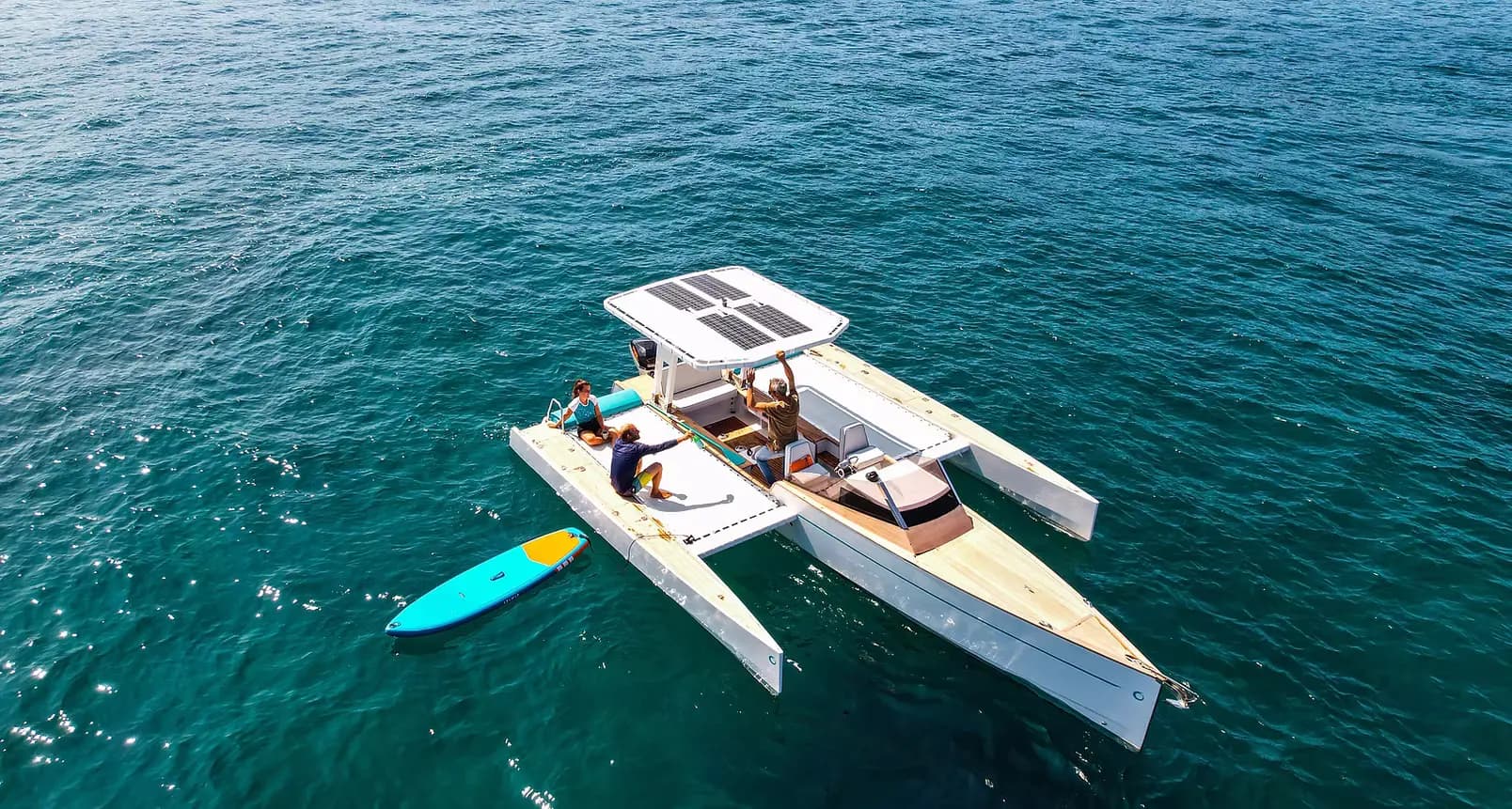 Novaluxe X-Wing catamaran viewed from above on open water, with a central cabin and blue paddleboard alongside