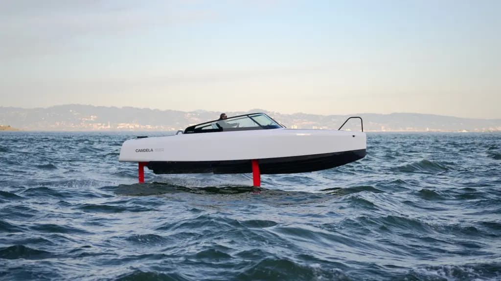 White Candela C-8 electric boat traveling above choppy water on submerged red hydrofoils, with a distant shoreline in the background.