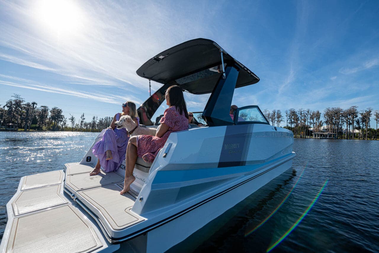 Side view of a blue wake boat (ingenity) from the stern, showing the swim platform, aft seating, tower, and two passengers on calm water.