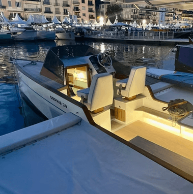 Novaluxe X-Wing electric boat moored in a marina, showing its open cockpit, twin cream seats, illuminated cabin, and waterfront surroundings.
