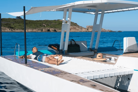 Novaluxe X-Wing electric boat with canopy and sunbather cruising near rocky coastline.