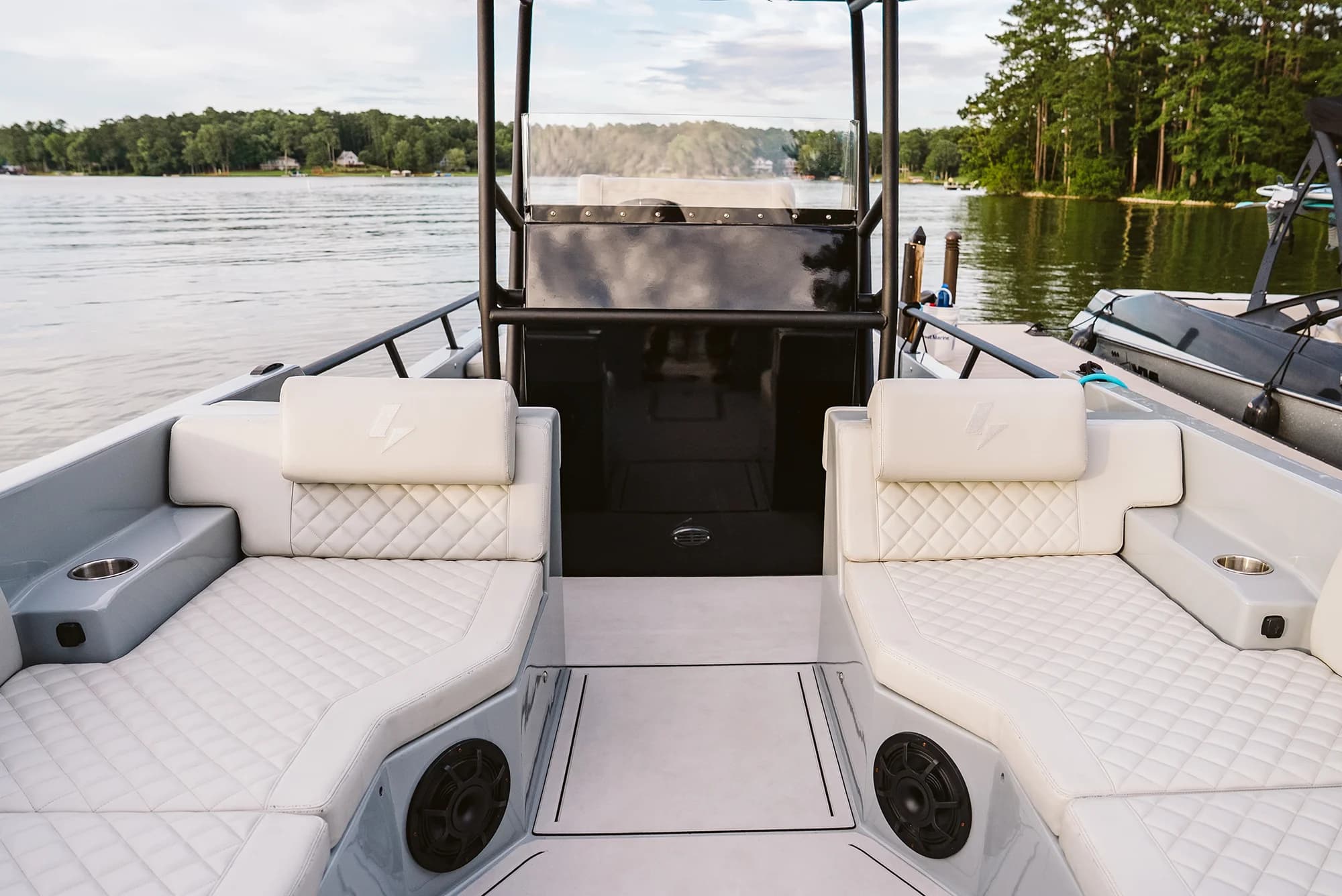 White open-deck electric boat (Halevai model2050) interior with quilted seating on sun lounges, center helm console, speakers, and a lake visible in background.