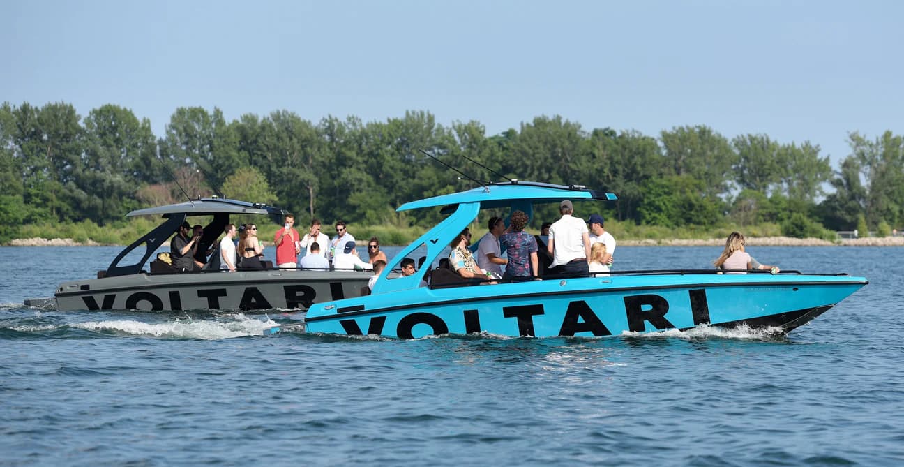 Two turquoise Voltari boats carrying passengers cruise side by side on a lake, with a tree-lined shoreline in the background.