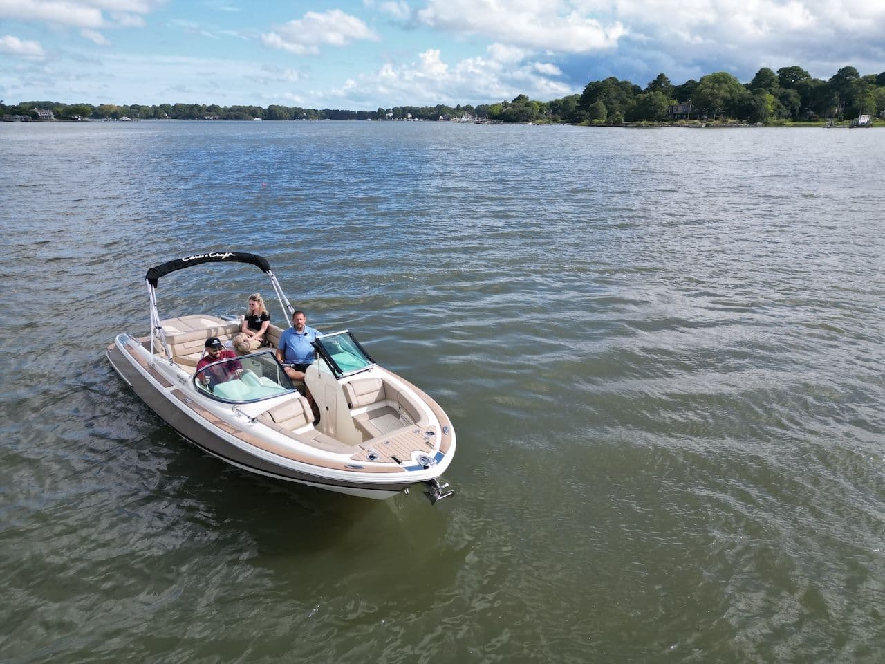 Chris-Craft bowrider with two passengers and a captain on calm water near a wooded shoreline.