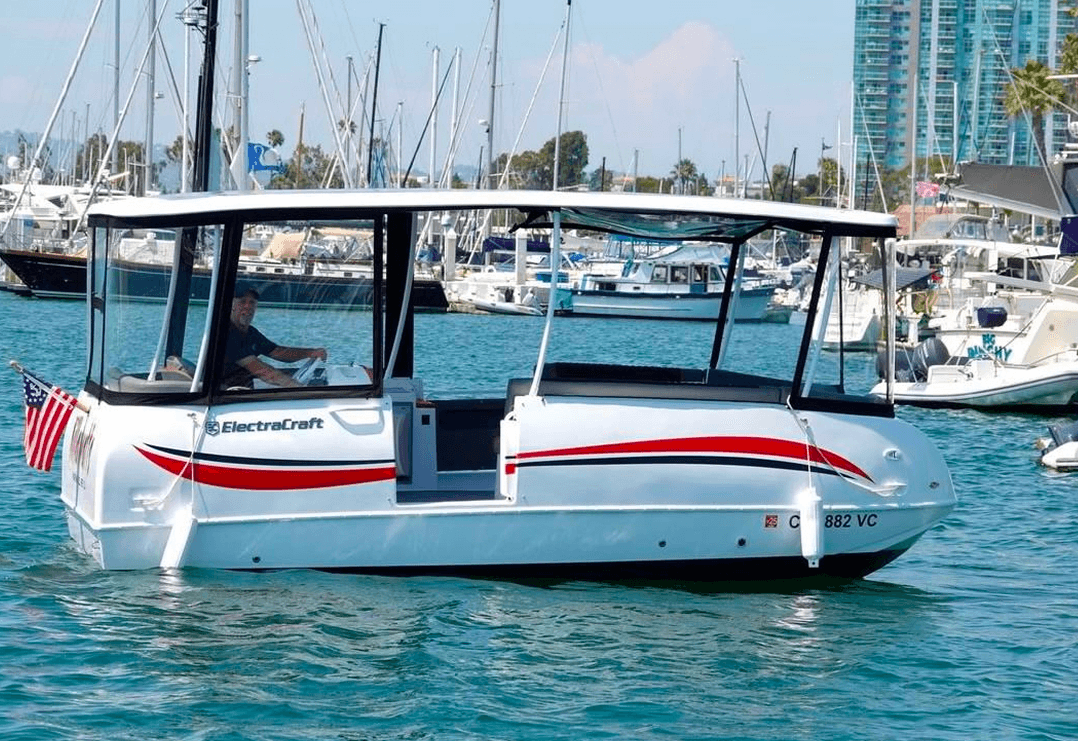 ElectraCraft 182TR electric boat in a marina, with a covered cabin, white hull with red accents, and yachts and waterfront buildings behind it.