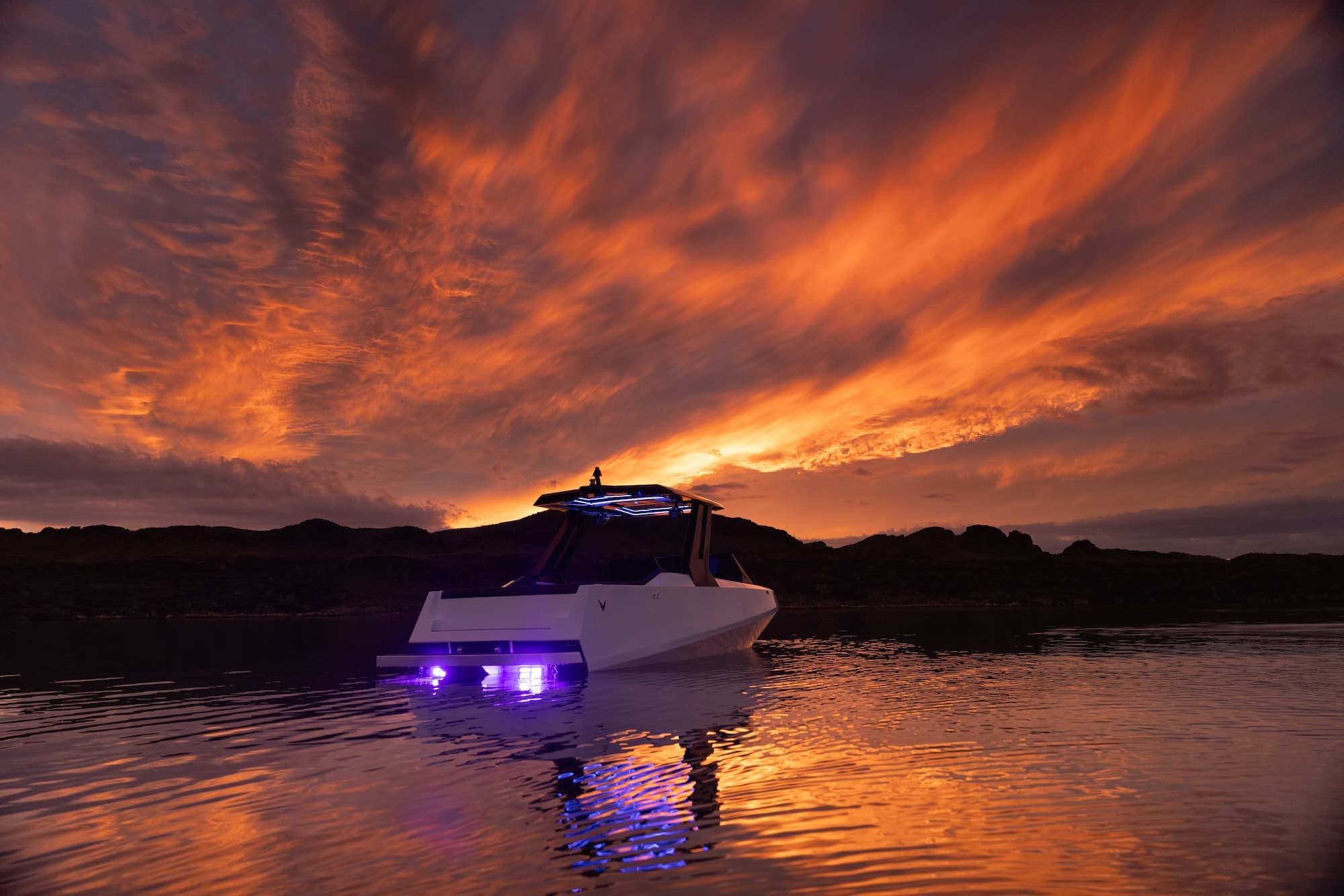 Voltaic AEW24 electric wakeboat cruising at dusk, with purple underwater lights reflecting on calm water beneath a dramatic orange sunset.