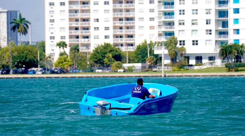 Blue electric motorboat carrying one passenger on turquoise water, with waterfront high-rise buildings and palm trees in the background.
