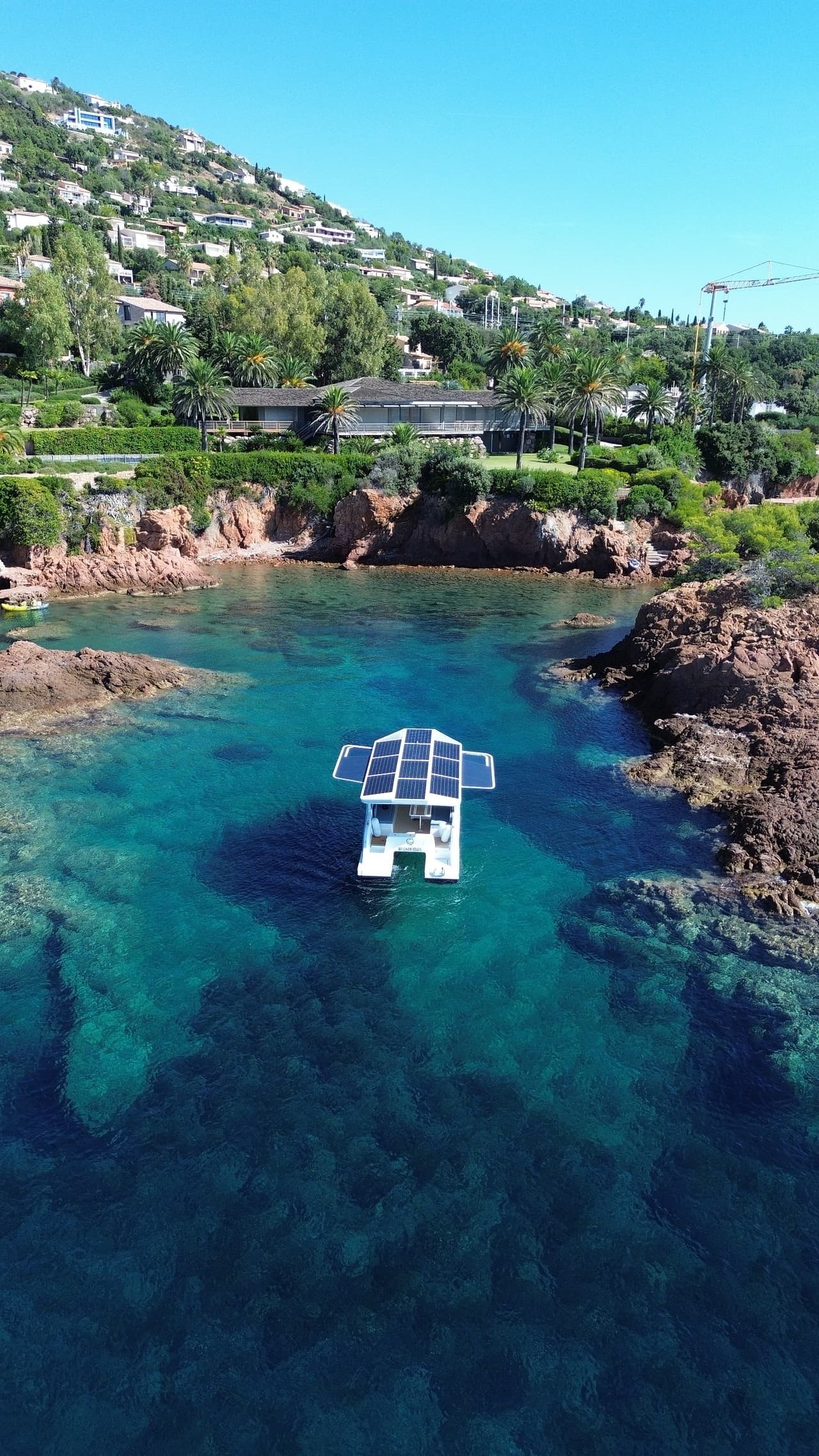 Catamaran cruising through clear turquoise water near a rocky coastline and hillside homes with solar panel roof and wings deployed