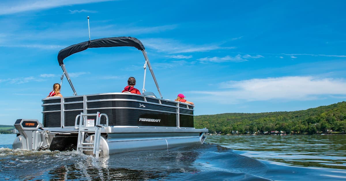 Princecraft Brio E17 pontoon boat with raised canopy and outboard motor cruising on a calm lake near a wooded shoreline