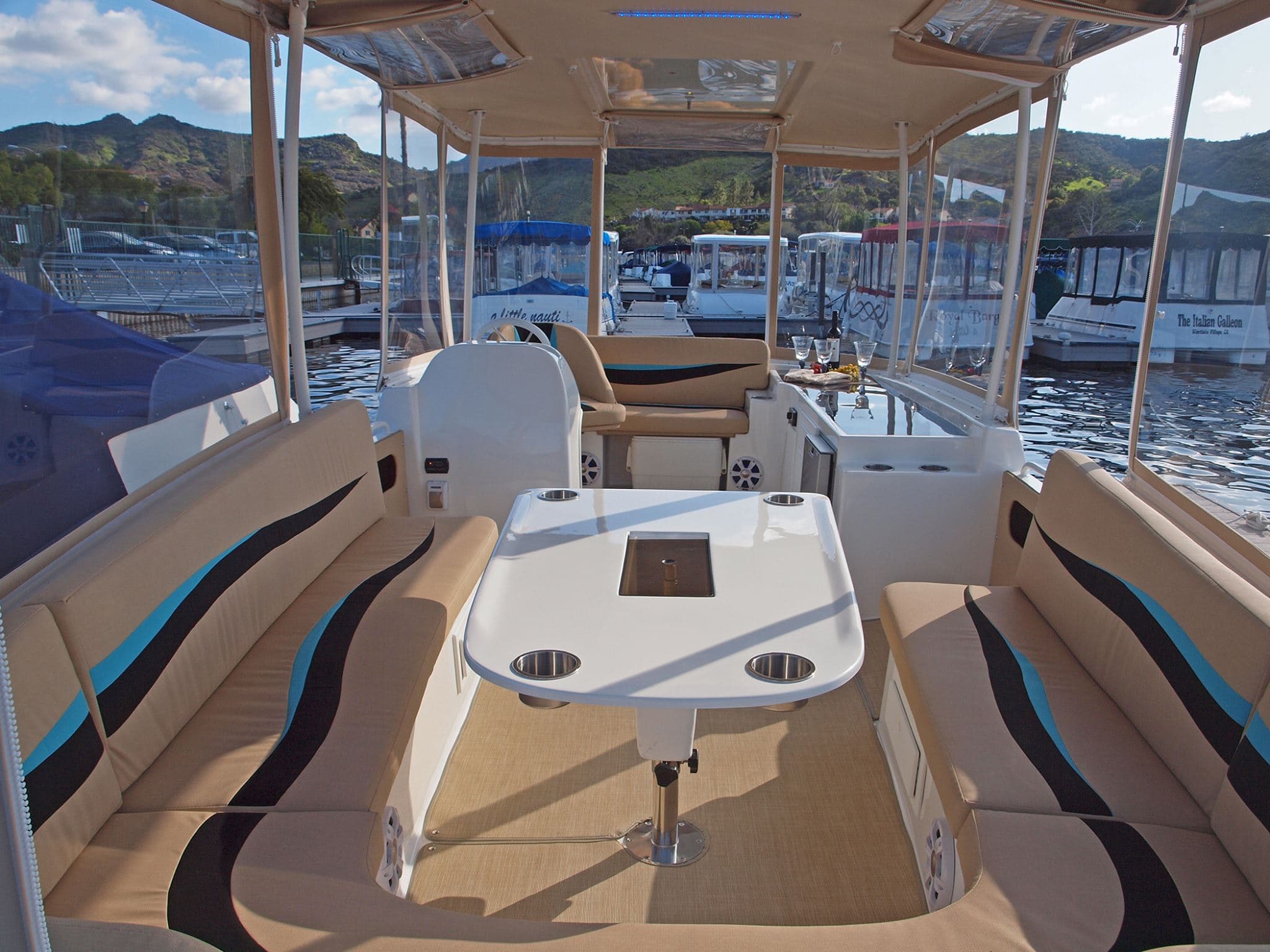 Interior of an ElectraCraft 182TR-style electric boat with wraparound tan and multi color accent seating, central table, helm, canopy, and marina views.