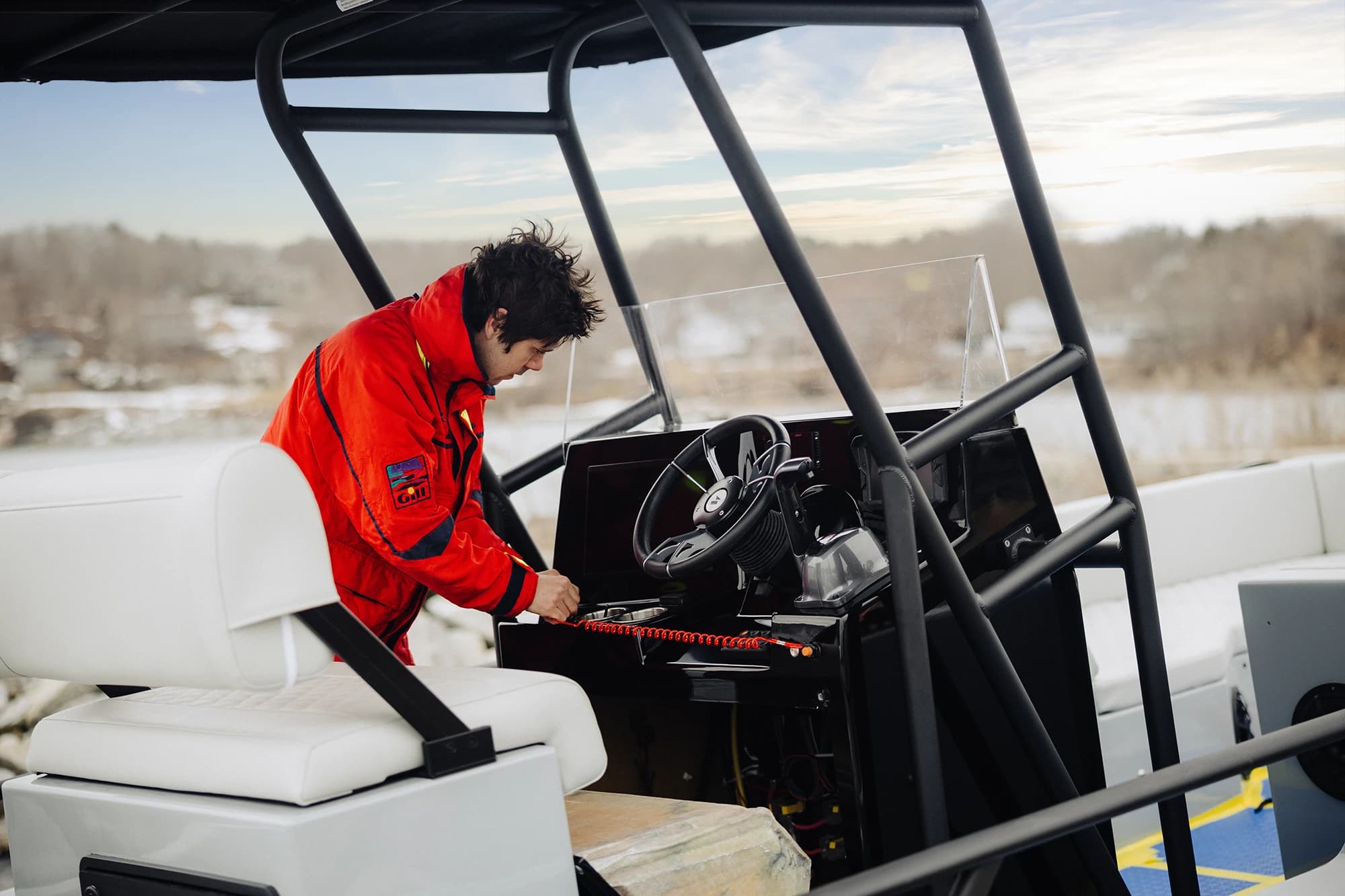 Person in a red jacket adjusts the controls of a Halevai model2050 electric boat on a calm, wintry lake.