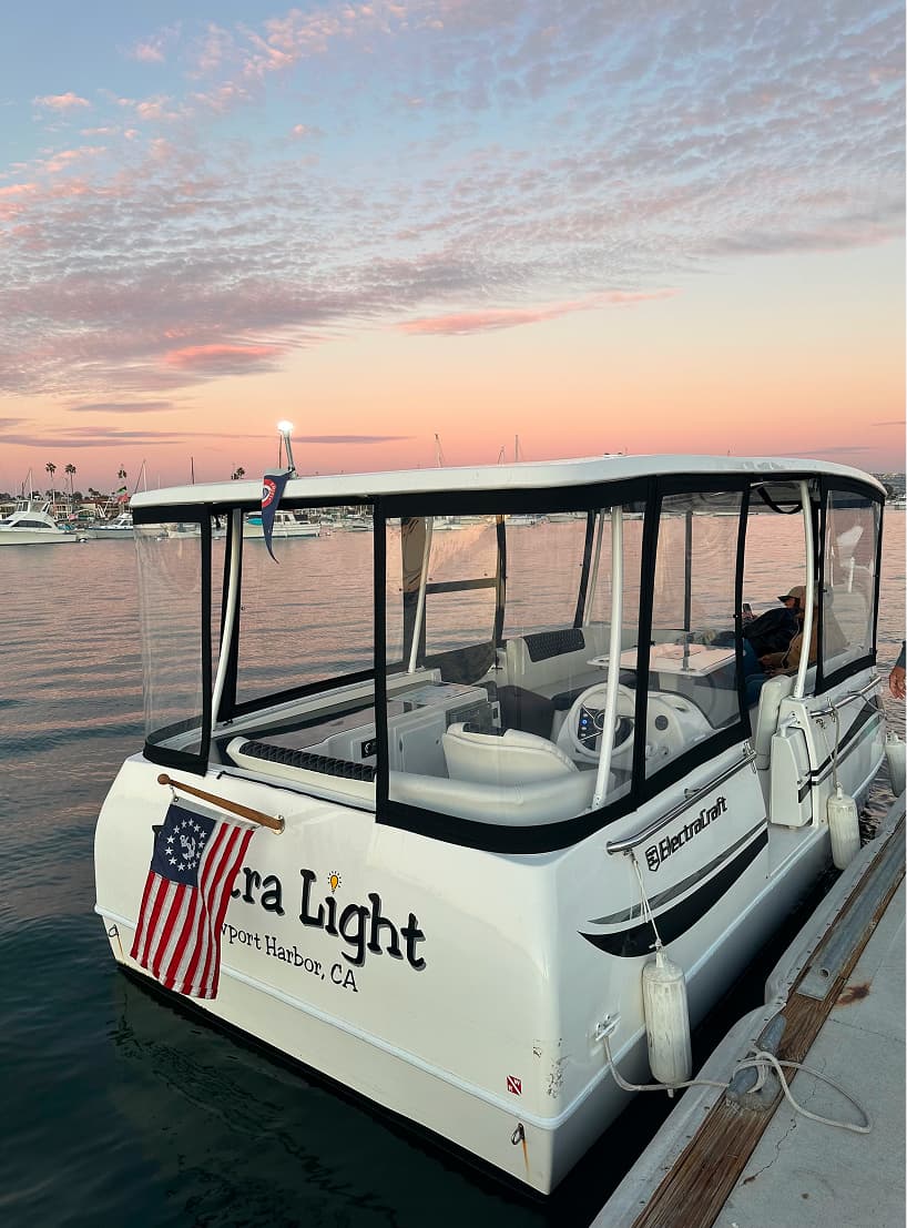ElectraCraft 182TR enclosed electric boat docked at sunset, with clear side panels, white seating, canopy, and American flag at the stern.
