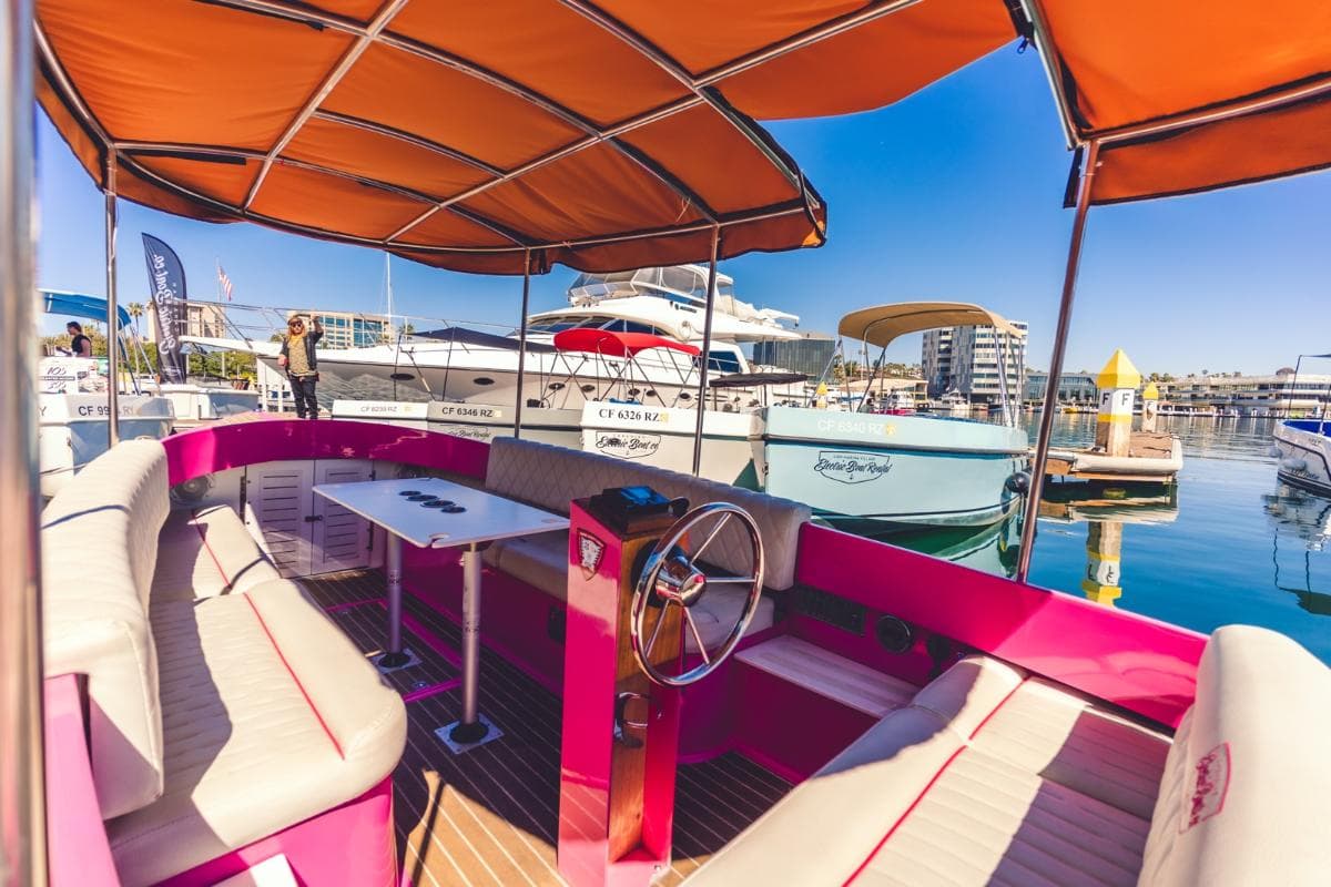 2024 Vision Marine Fantail electric boat cockpit with pink-and-cream seating, wood helm, orange canopy, and marina backdrop.