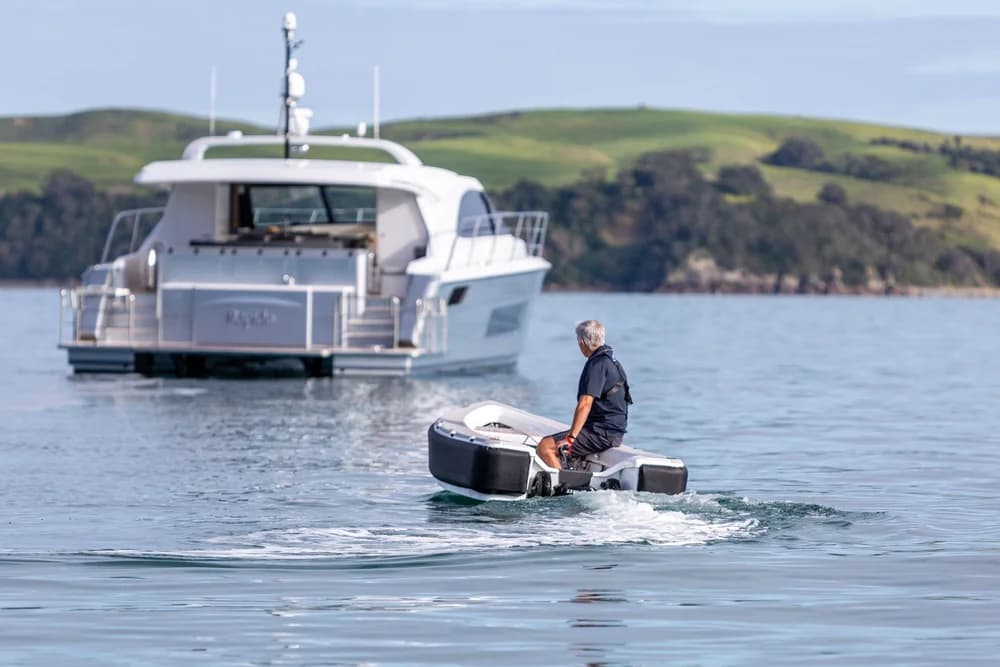 Ocean Cruising Tenders OCT 300 tender cruising in the foreground, with a larger motor yacht anchored near a green, rocky shoreline.