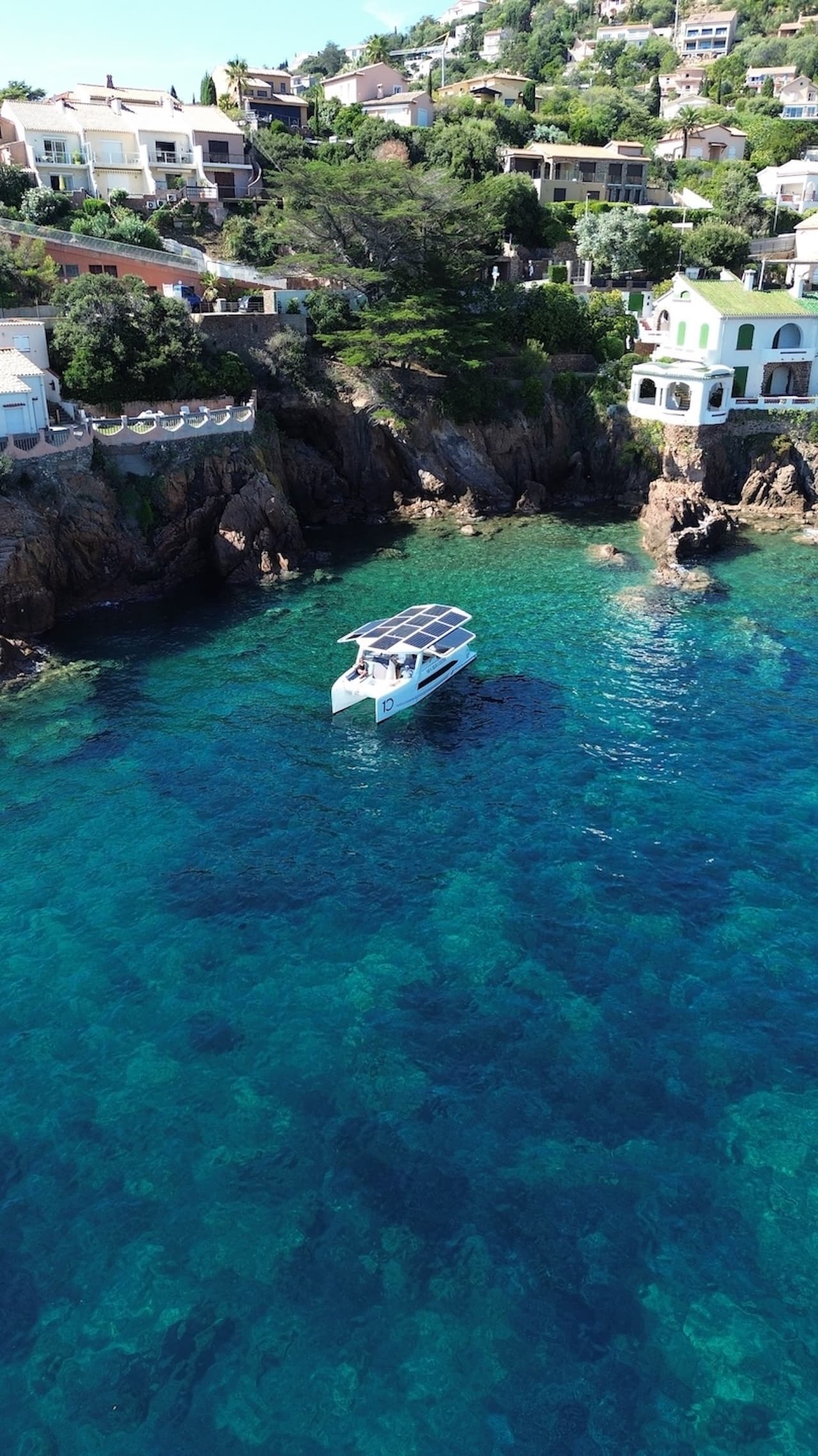 Millikan M10 viewed from above, cruising in clear turquoise water near a rocky coastline and hillside homes