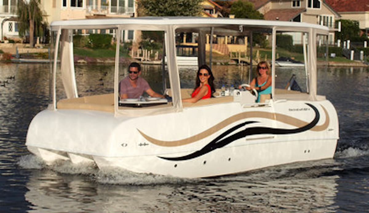ElectraCraft 162TR electric pontoon boat cruising on calm water, with three passengers aboard and waterfront homes in the background.