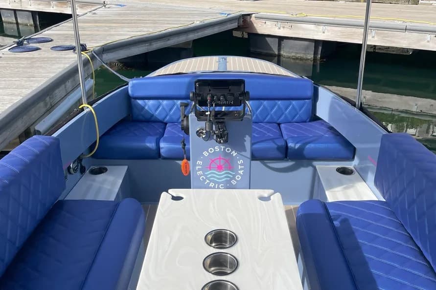 Interior of a blue electric boat (2024 Vision Marine Fantail 217) with wraparound seating, center console, white dining table, and docked waterfront surroundings.