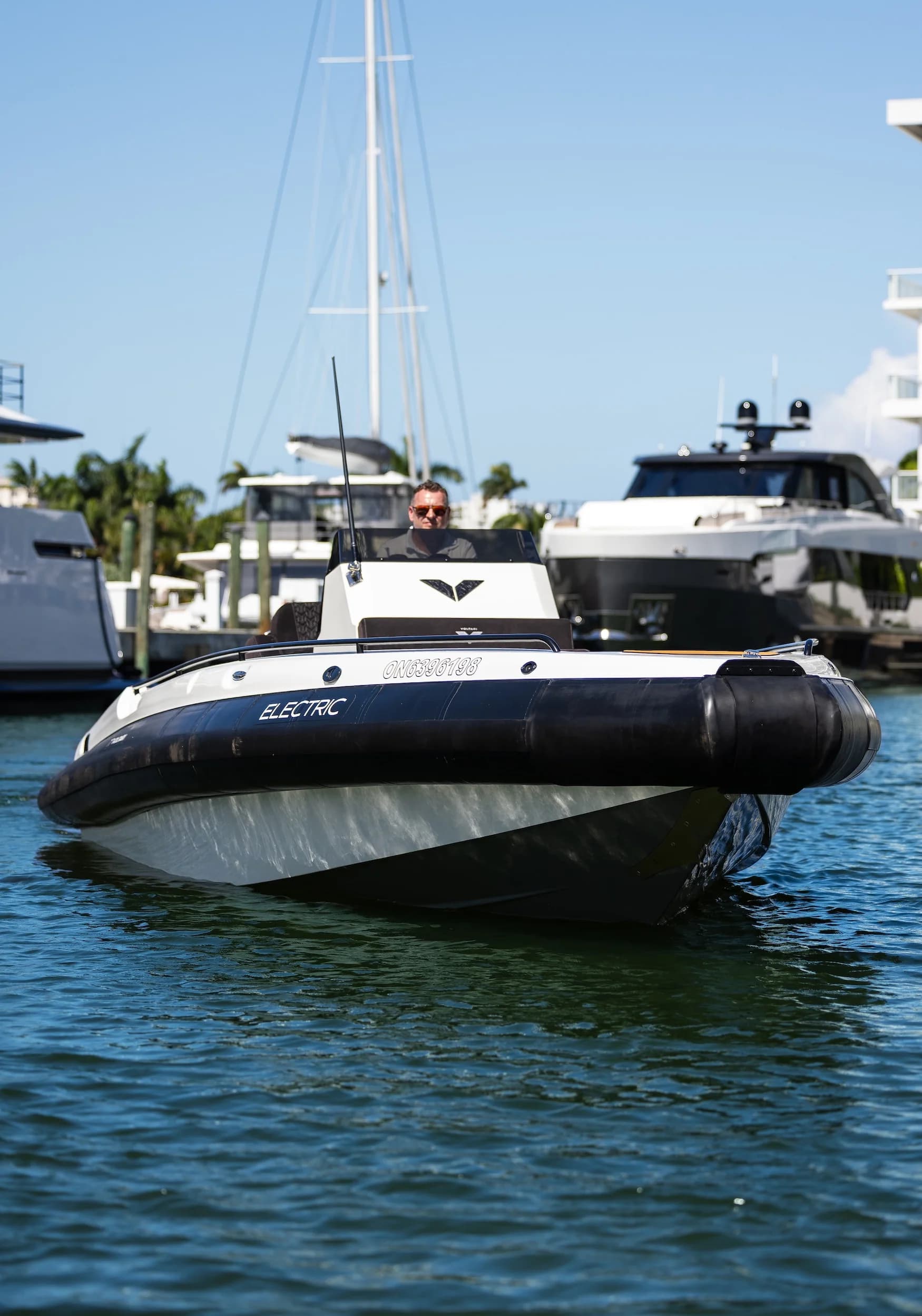 Voltari 260 T/T electric boat cruising on calm marina water, viewed from the front with yachts and palm trees in the background.