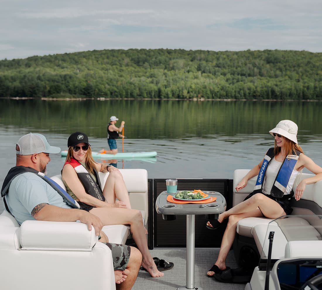 View from the stern into the cockpit of a pontoon boat (2026 Princecraft Vectra 21 RL) with three passengers, lounge seating, and a table on a calm lake.