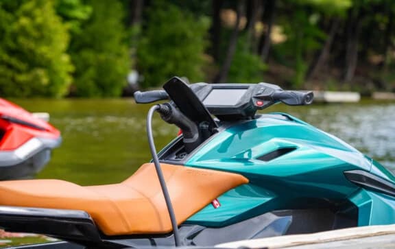 Teal Taiga Orca Performance electric personal watercraft with orange seat, parked on a calm, tree-lined lake.