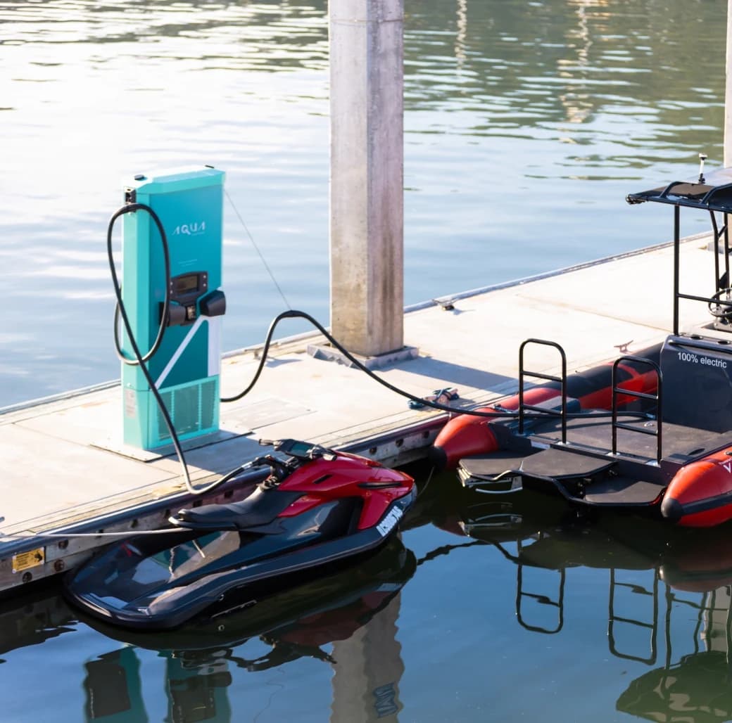 Electric personal watercraft (Taiga Orca P2) docked beside a turquoise charging station at a marina, with a second red-and-black watercraft partially visible.