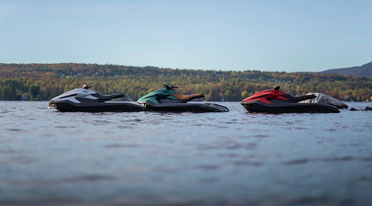 Three personal watercraft, including a Taiga Orca Performance, lined up on a lake with forested hills in the background.