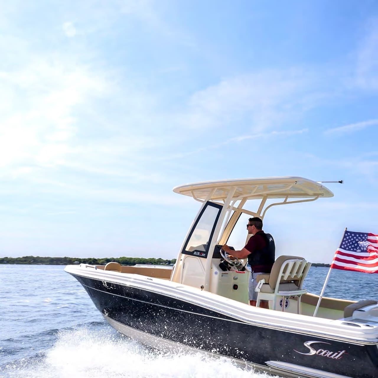 Scout 215 XSF center-console boat underway, viewed from the starboard side, with a T-top, American flag, and driver visible.