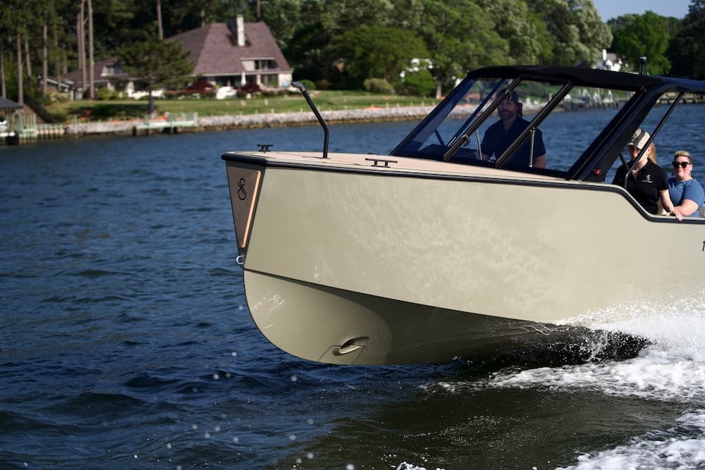 Front starboard view of a cream-colored X Shore 1 underway, with two visible passengers and water splashing along the bow.