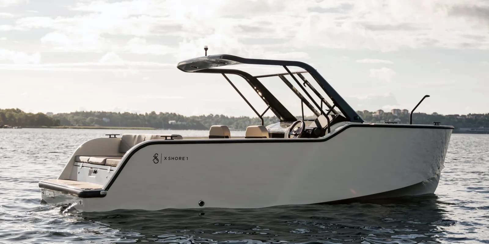 Side view of a white X Shore 1 motorboat on calm water, showing the hull, open stern platform, windshield, and black canopy frame.