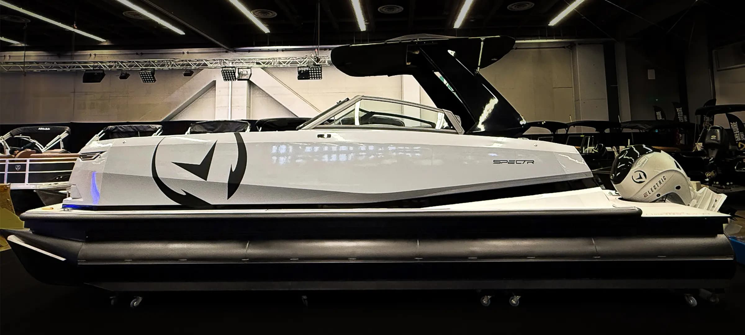 Side view of a white pontoon boat (2026 Vision Marine SPECTR) with a dark canopy and outboard motor displayed indoors.