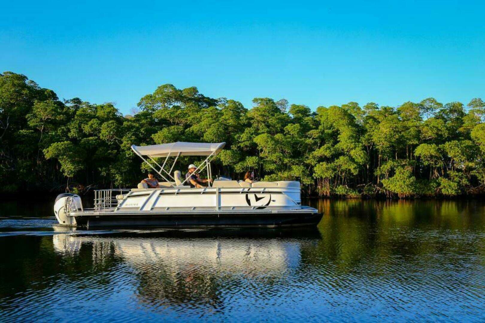 Side view of a pontoon boat (Vision Marine V24) cruising on a calm, tree-lined waterway