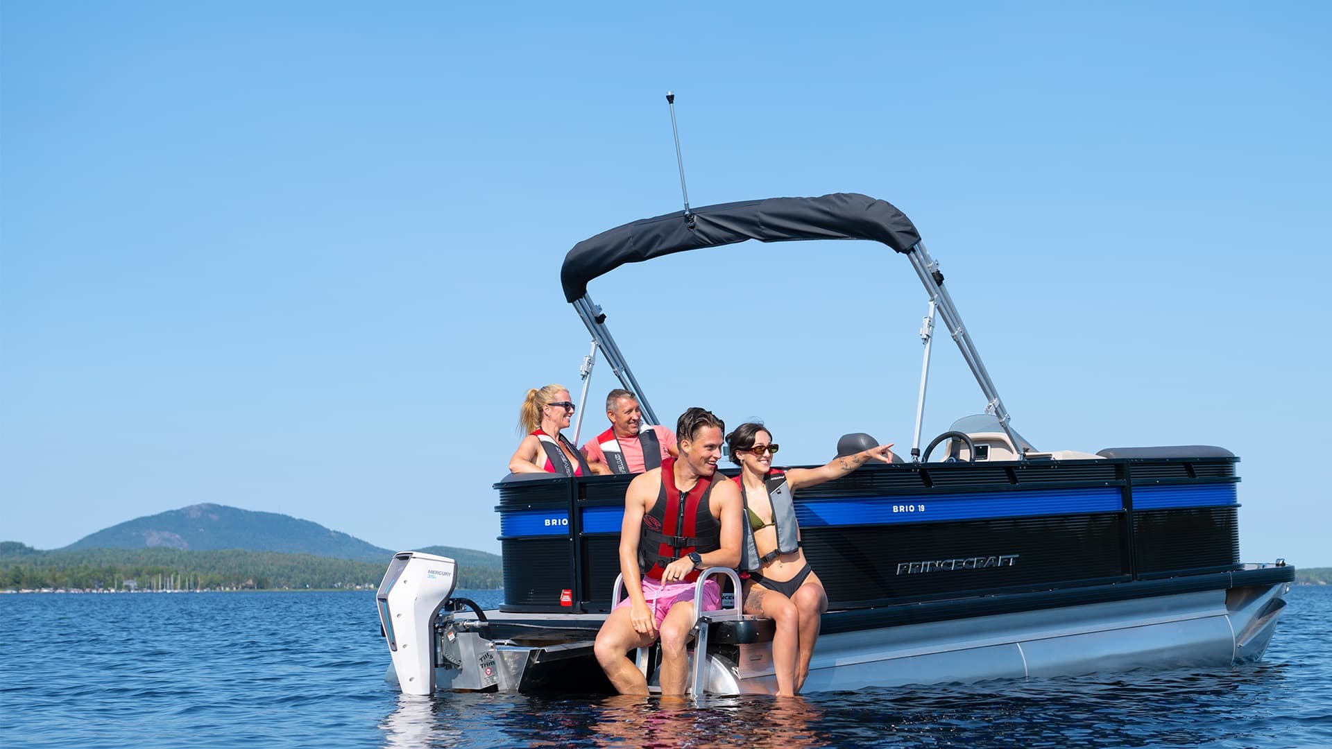 Princecraft Brio e 17 pontoon boat viewed from the side on a lake, with an outboard motor and several people seated at the stern.