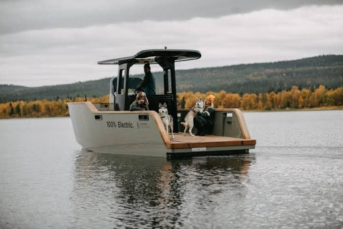X Shore Eelex boat viewed from the stern on a lake, with passengers and a dog aboard; “100% Electric” marking visible on the hull