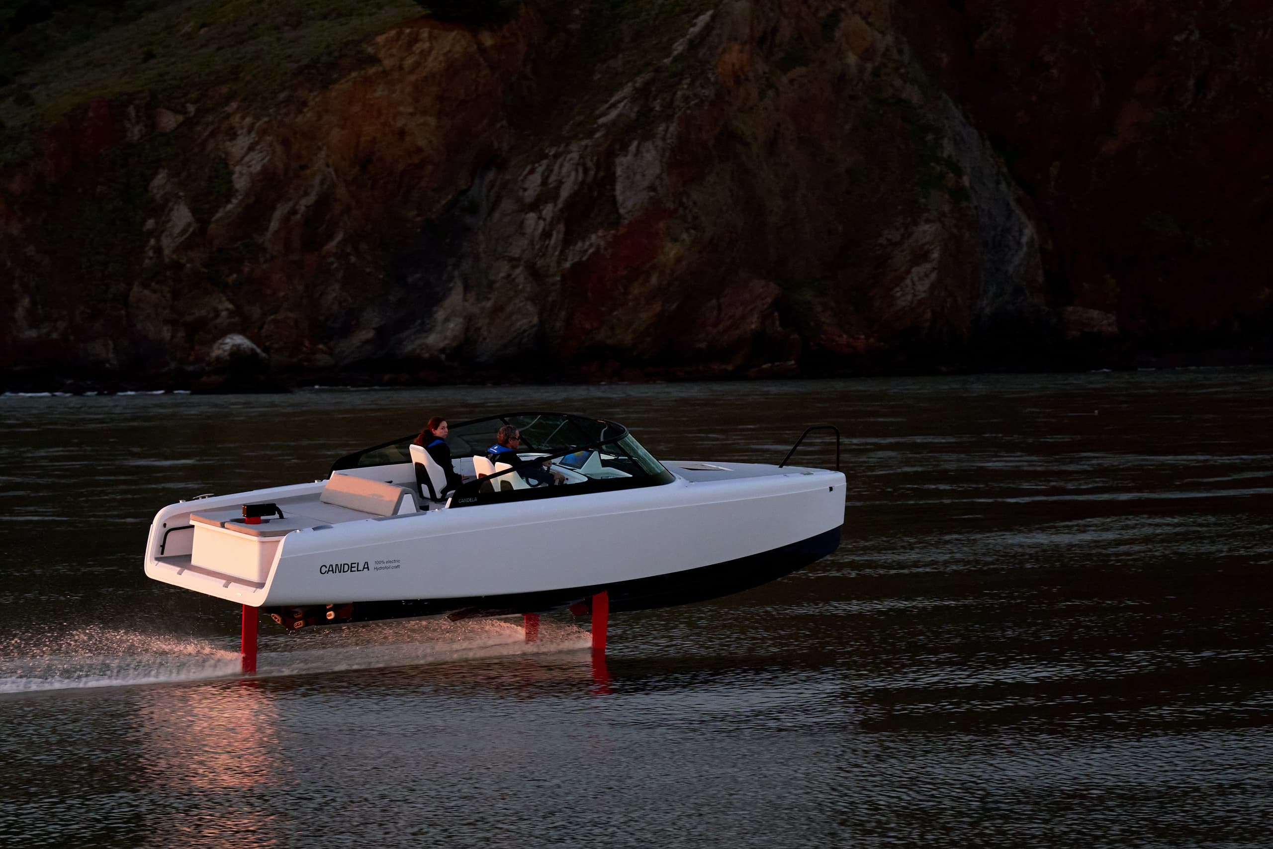 Candela C-8 hydrofoiling on a dark body of water near a rocky cliff, viewed from the side.