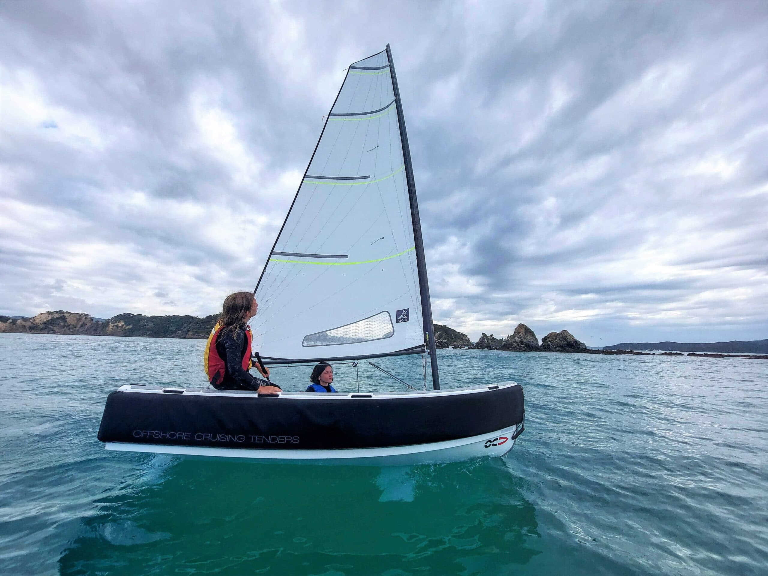 Offshore Cruising Tenders OC330 sailing on coastal waters, viewed from the side with its raised sail and two occupants visible.