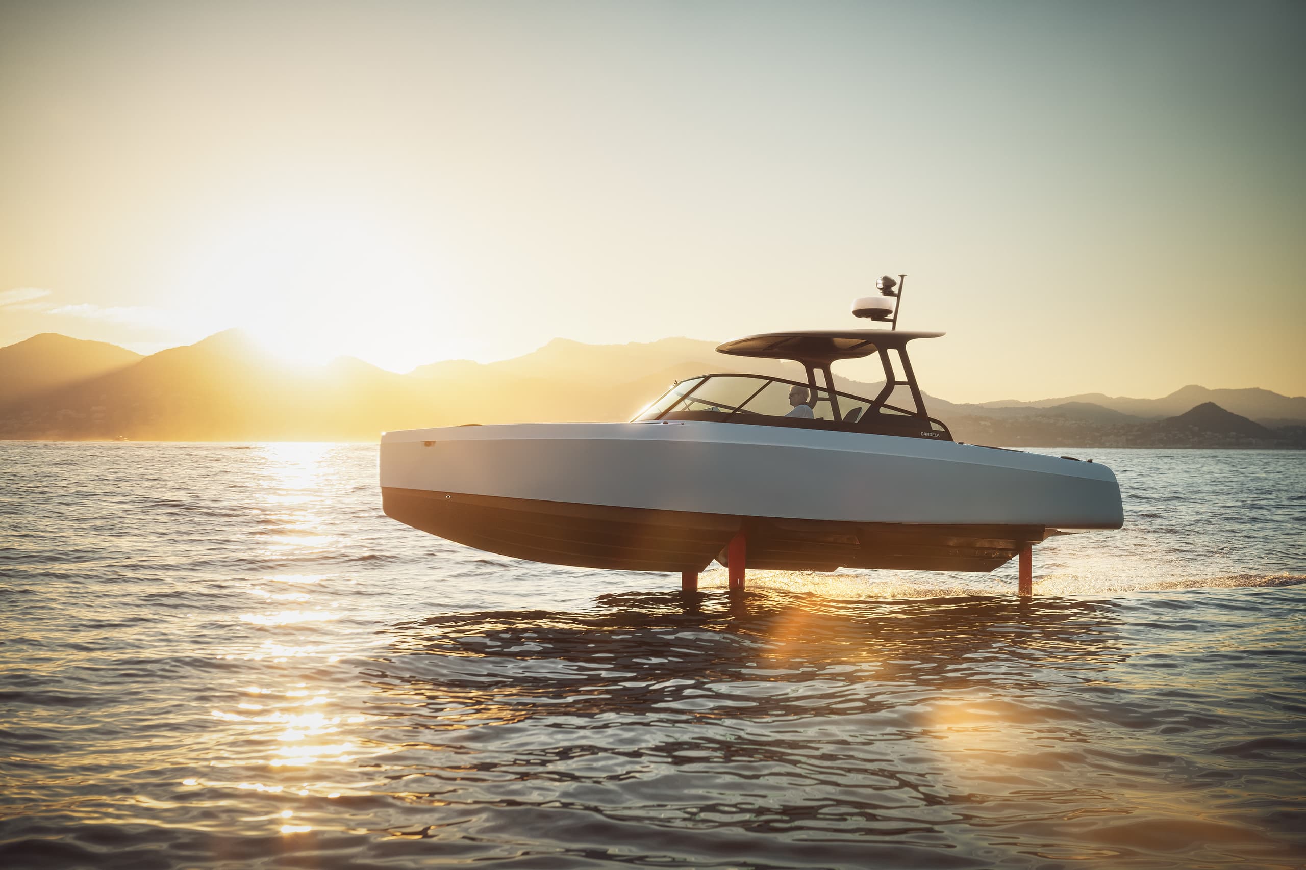 Candela C-8 cruising on hydrofoils at sunset, viewed broadside on calm water with mountains in the background