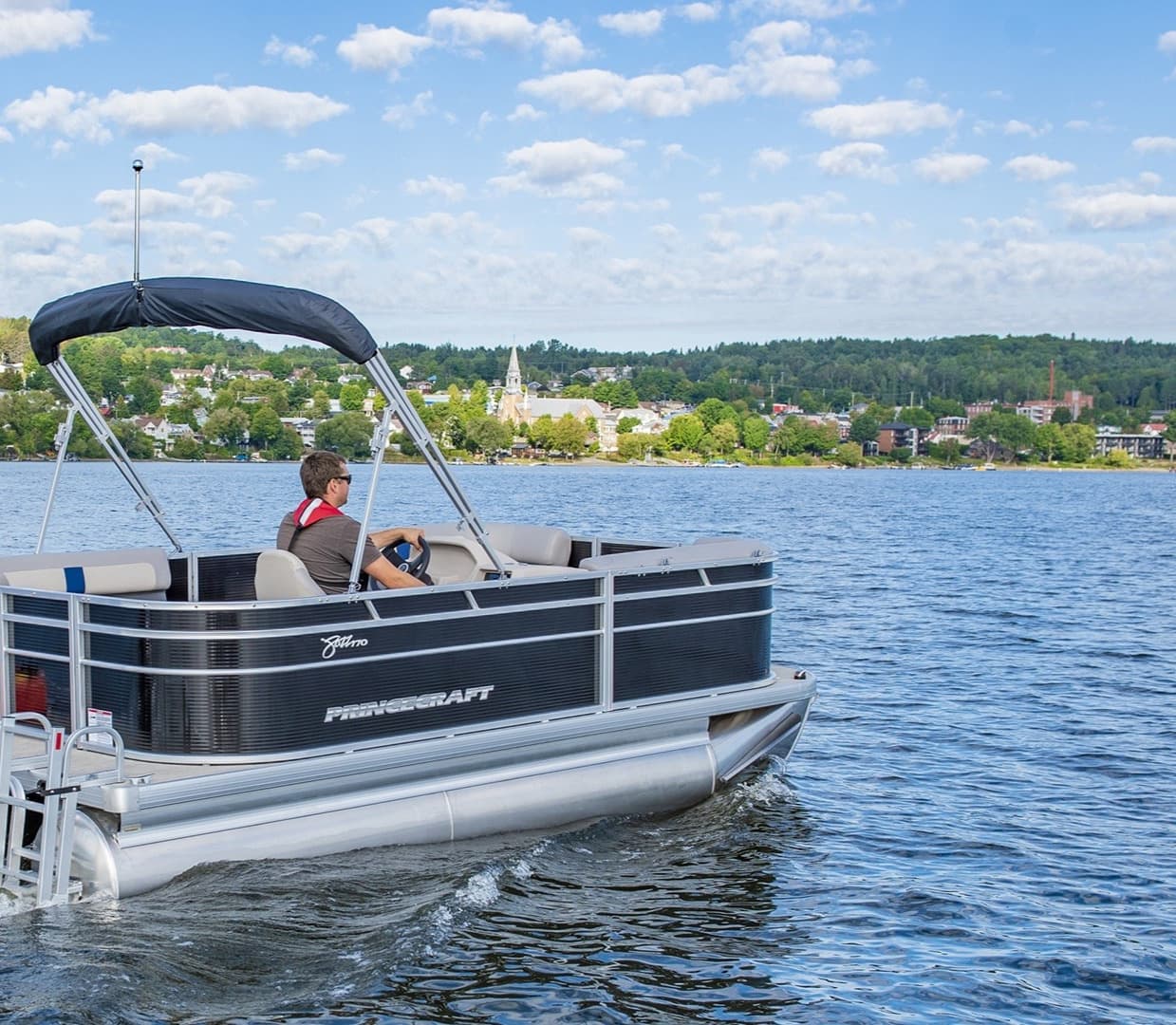 Princecraft Jazz 17 pontoon boat viewed from the side on a lake, with a canopy and person seated at the helm.