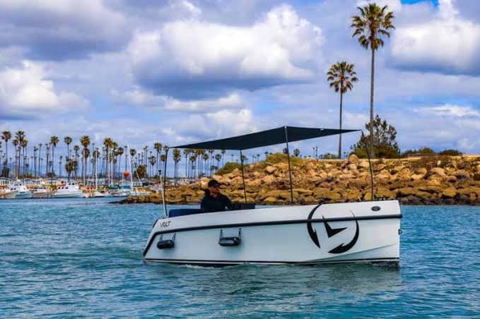 Vision Marine Volt 180 viewed from the side on the water, with marina, breakwater, palm trees, and canopy visible.