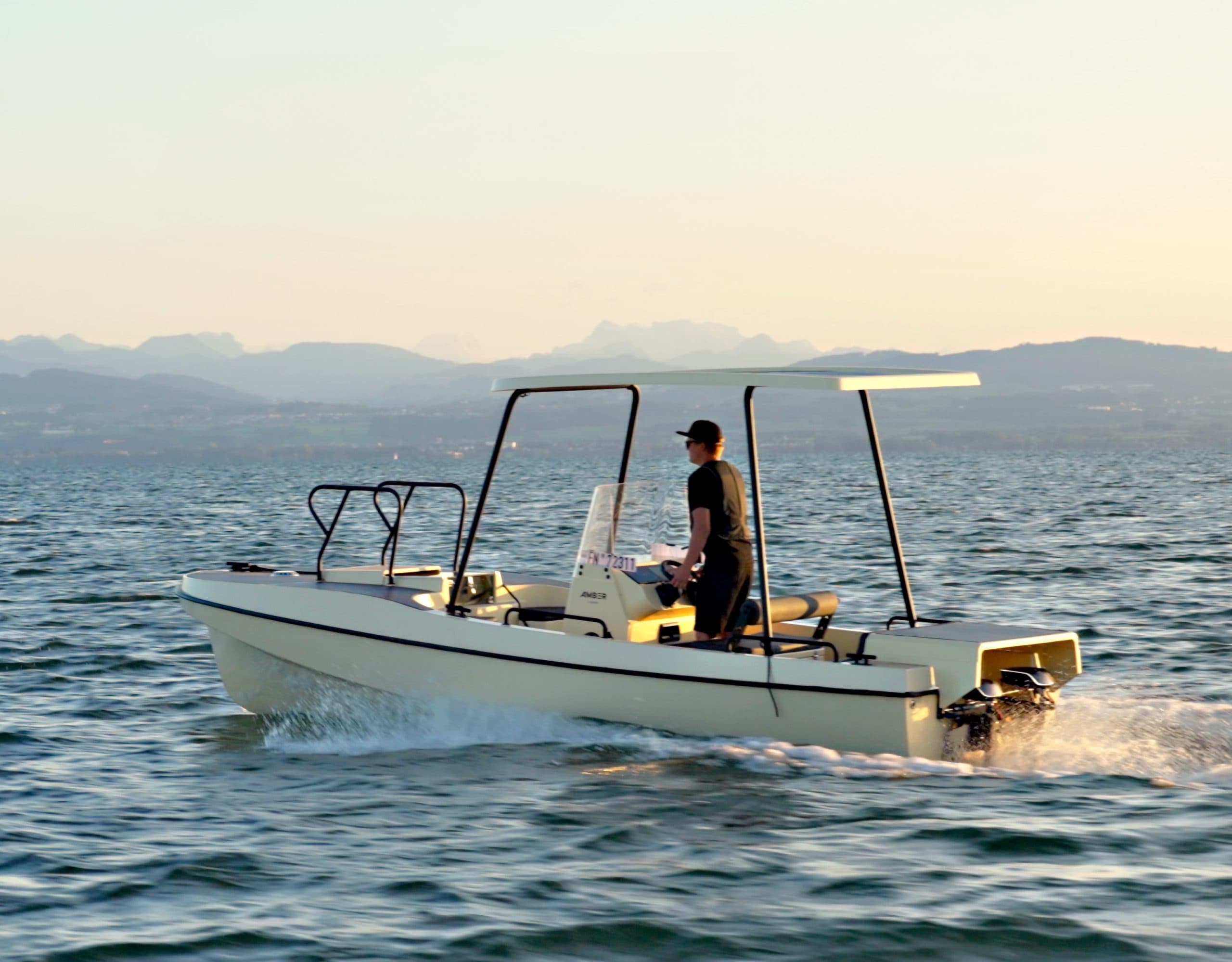 Elvene Amber electric boat cruising on open water with a standing operator, canopy, and mountainous shoreline in the background.