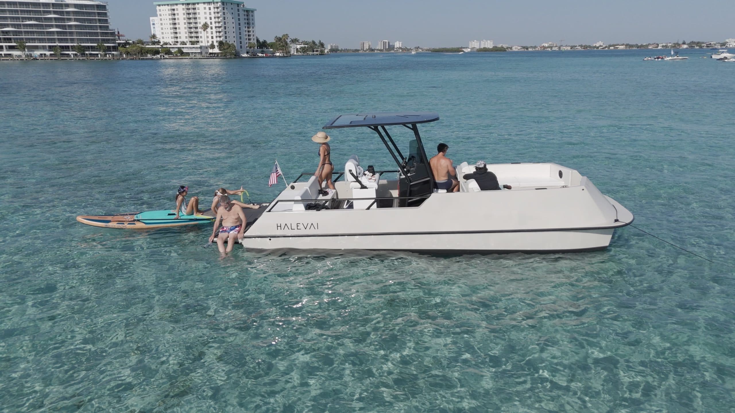 Halevai model2050 electric boat cruising in clear shallow water, with people aboard and swimmers alongside, near a coastal city.