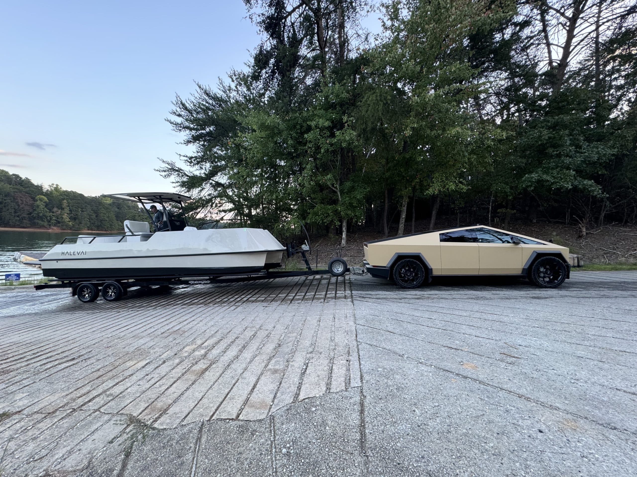 Electric boat (Halevai model2050) with canopy on a trailer, hitched to a beige Cyber Truck vehicle beside a lake and wooded shoreline.