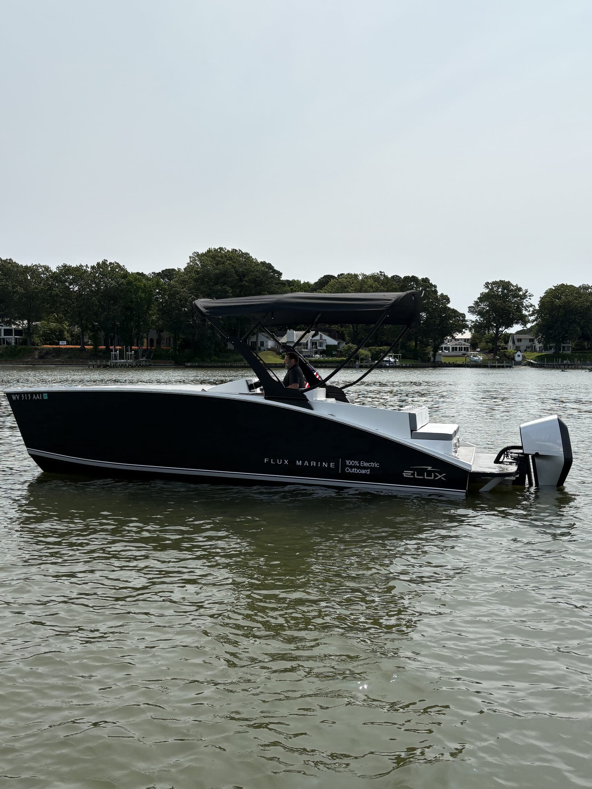 Black Flux Marine GFX Stealth boat with canopy and outboard motor on calm water near a tree-lined shore.