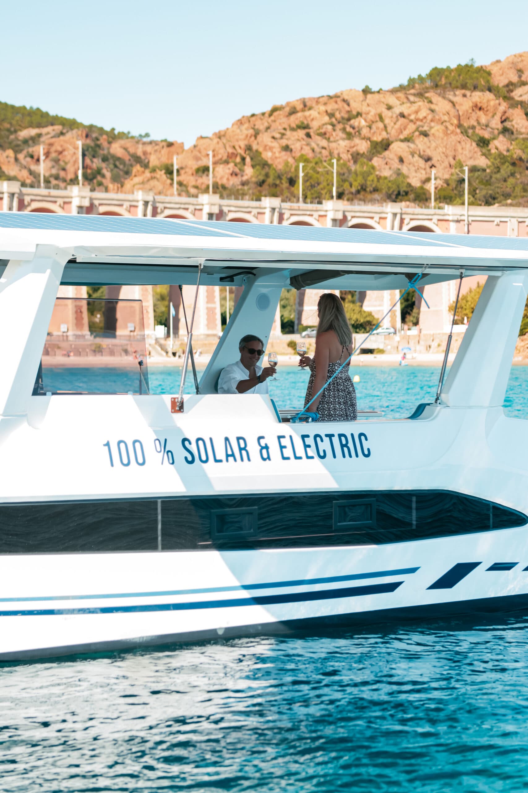 Millikan M10 side view on the water, with two people visible beneath the canopy and “100% solar & electric” lettering on the hull
