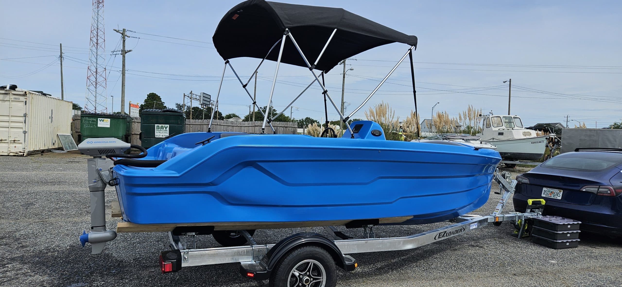 Blue Vision Marine Phantom electric boat on a trailer, fitted with a black canopy and hitched to a car in an outdoor lot.