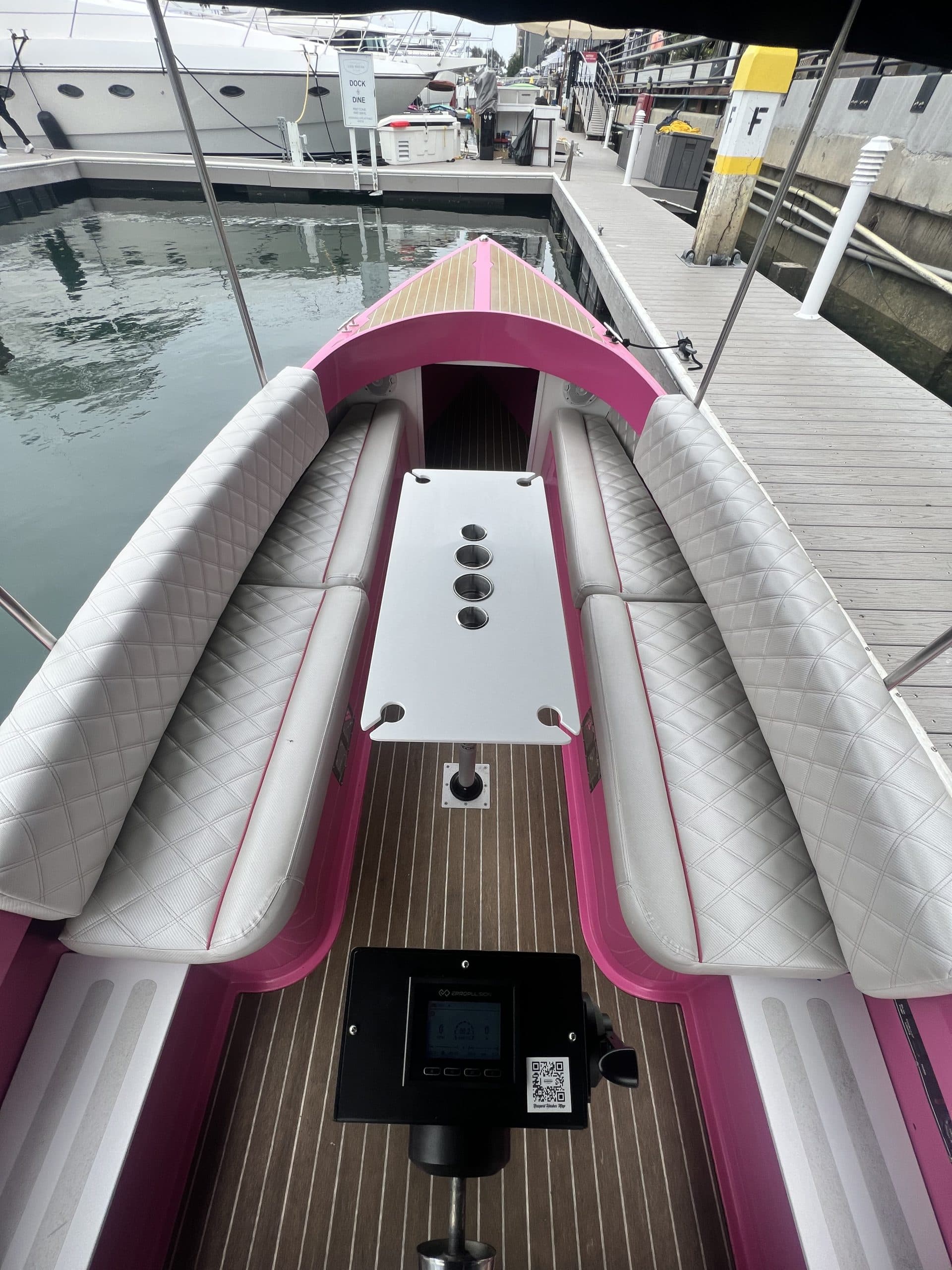 2024 Vision Marine Fantail cockpit with pink trim, white cushioned seating, wood-look flooring, helm, and marina dock surroundings