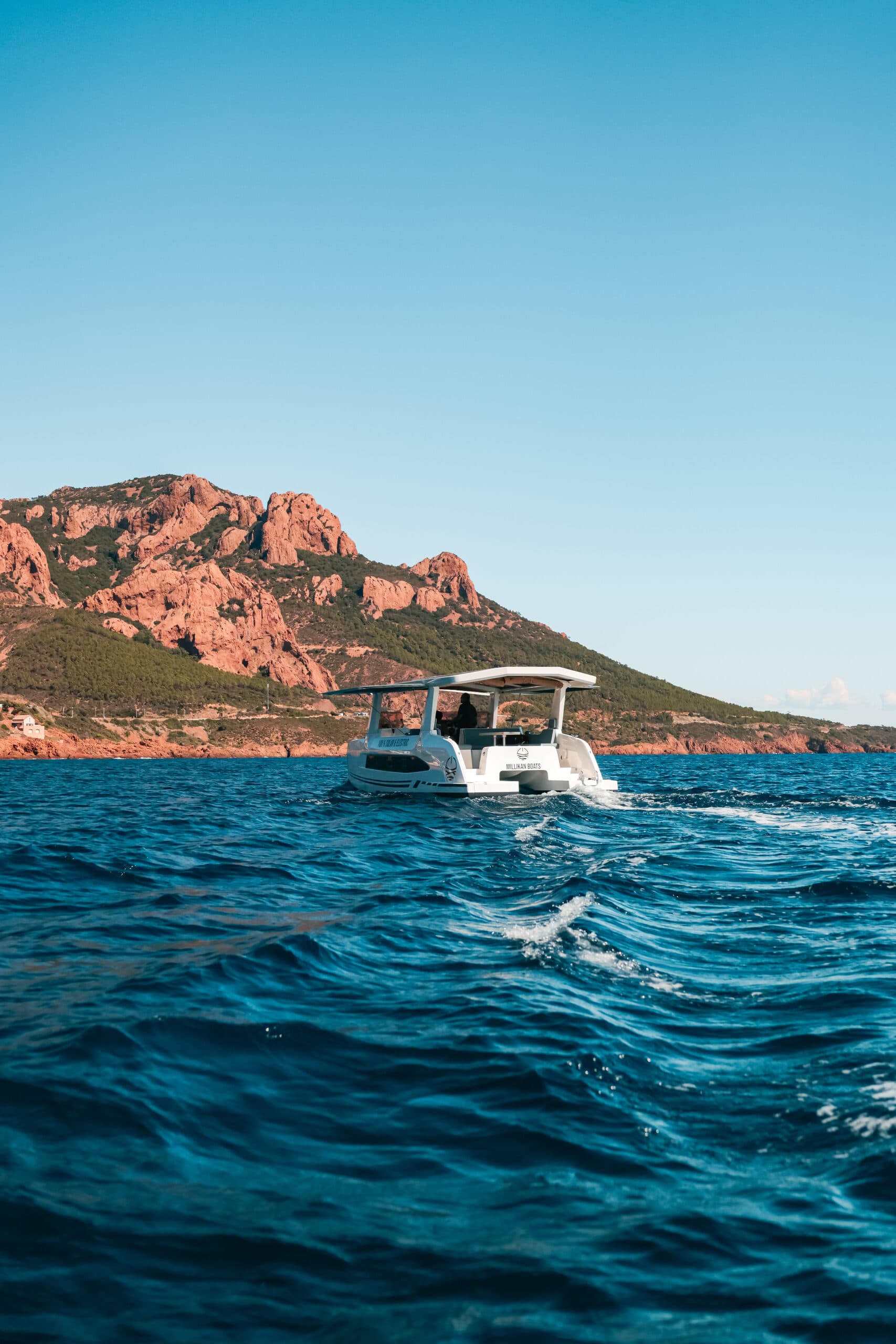 White electric catamaran (Millikan M10) cruising on blue coastal waters with rugged reddish mountains in the background with wings deployed on side